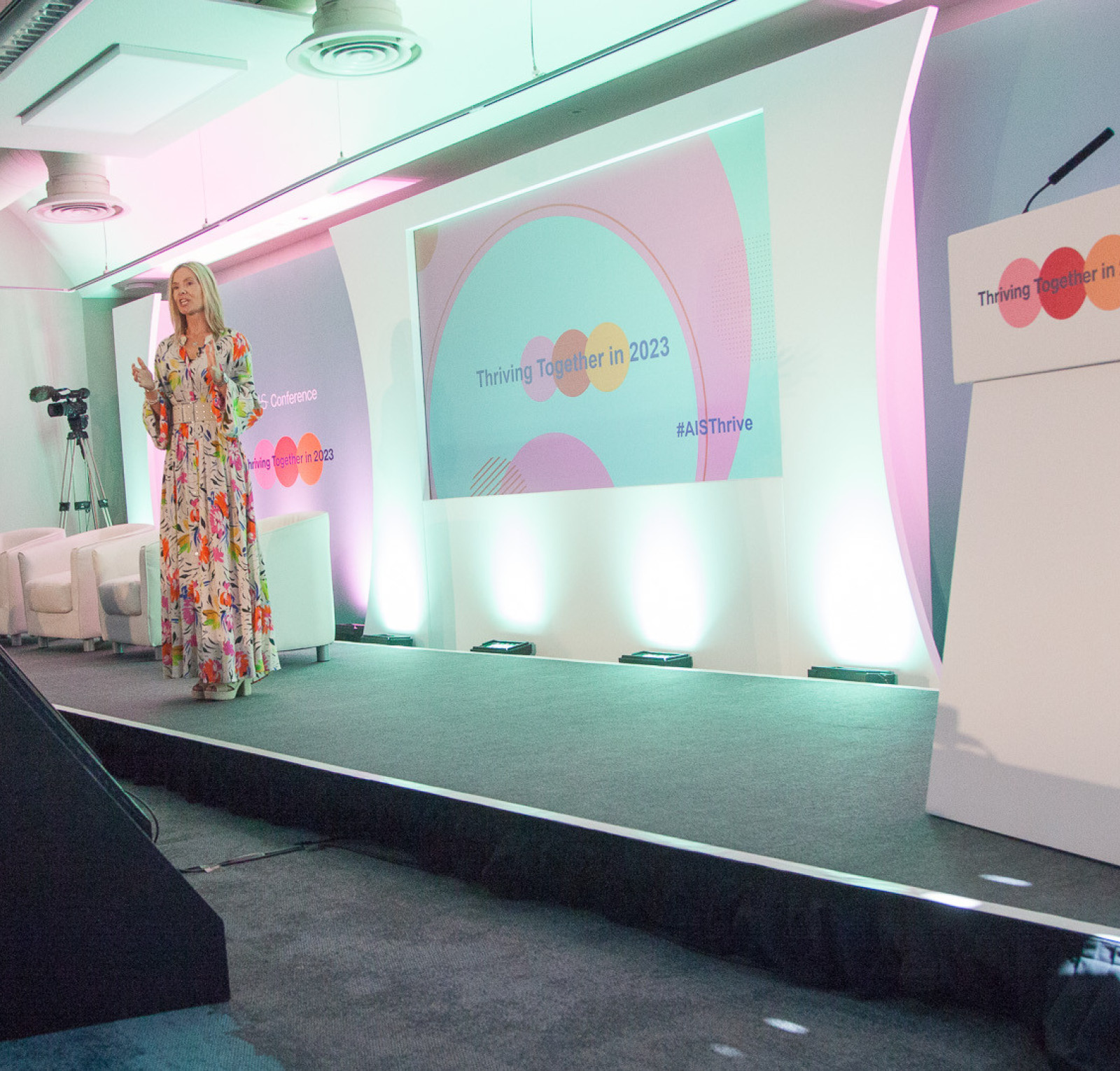 A speaker stands on stage at a conference, wearing a colorful floral dress. Behind her is a large screen displaying the event title 