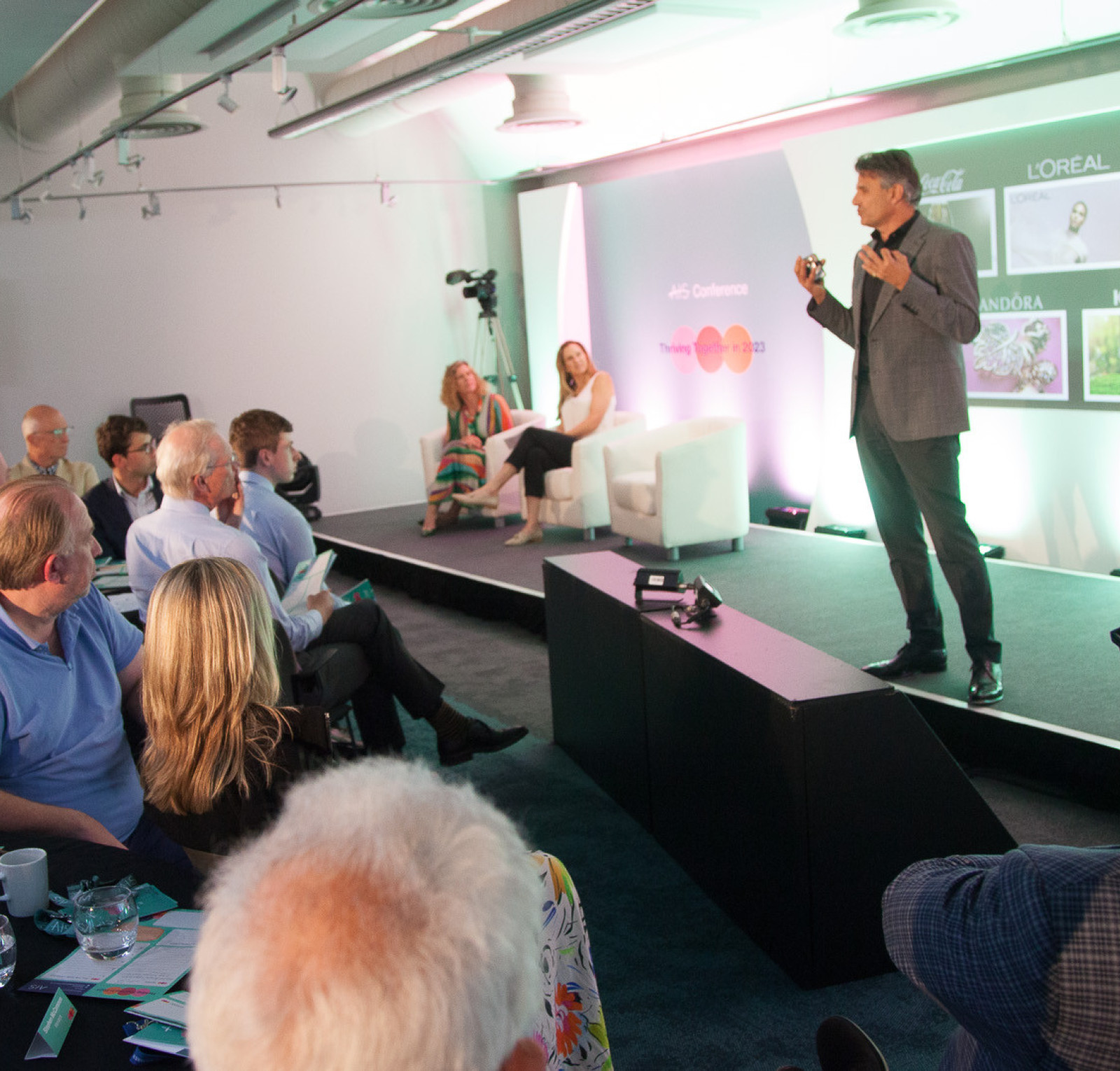A speaker presents on stage at a conference, with a screen displaying logos of L’Oréal, The Body Shop, and Kantar. Attendees are seated at tables, engaged in the discussion.