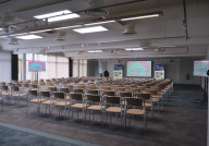 A spacious conference room set up with rows of chairs facing a stage. Two large screens display colorful presentations. Sunlight filters through large windows, illuminating the neutral-toned decor.