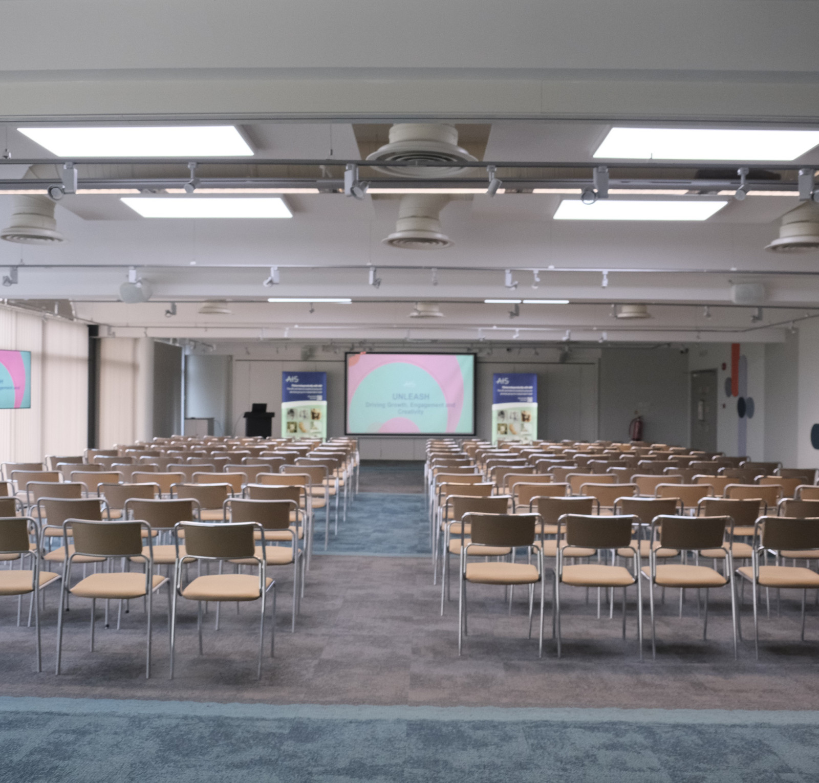 A spacious conference room set up with rows of beige chairs facing a stage. Two large screens at the front display pastel-colored presentation slides. Natural light fills the room through large windows, and the floor alternates between carpet and tiles.