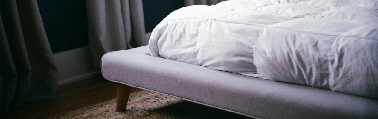 A corner of a cozy bedroom featuring a gray upholstered bed with a white comforter and a textured area rug on wooden flooring. Soft curtains frame a window in the background.