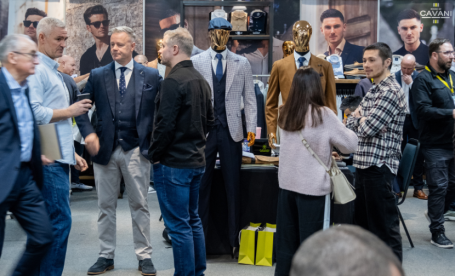 A group of men in suits and casual attire engage in conversation at a trade event. Mannequins dressed in stylish outfits are displayed on tables, with various accessories in the background. Attendees mingle in a bustling atmosphere.