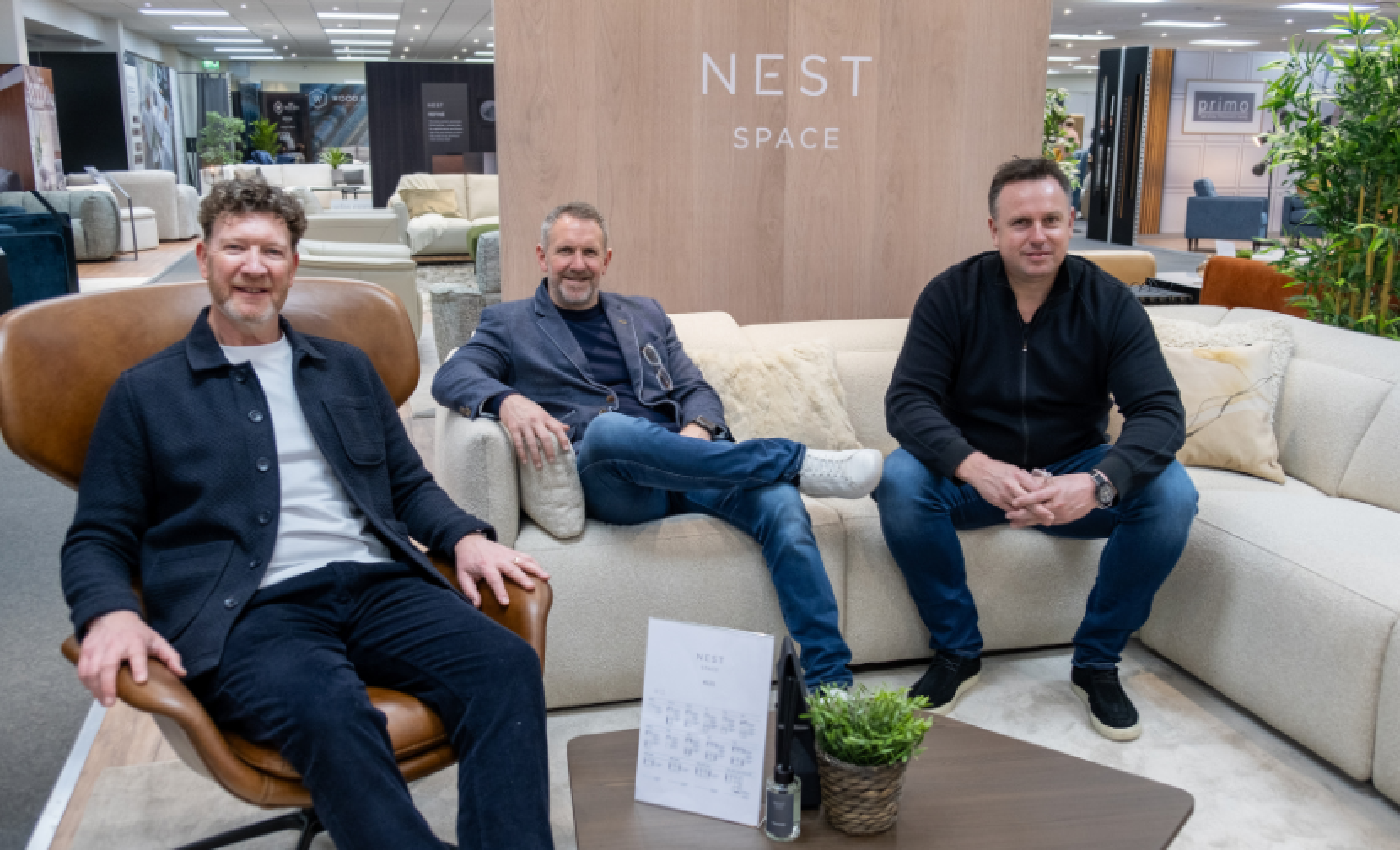 Three men seated on a large sofa in a modern showroom. The backdrop features a wooden wall with the words 
