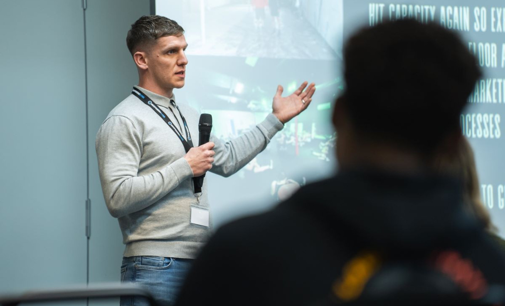 A man with short hair and a gray sweater is speaking into a microphone while gesturing with his right hand. He appears to be presenting in front of an audience. In the background, a slide displays images related to a fitness or training facility.