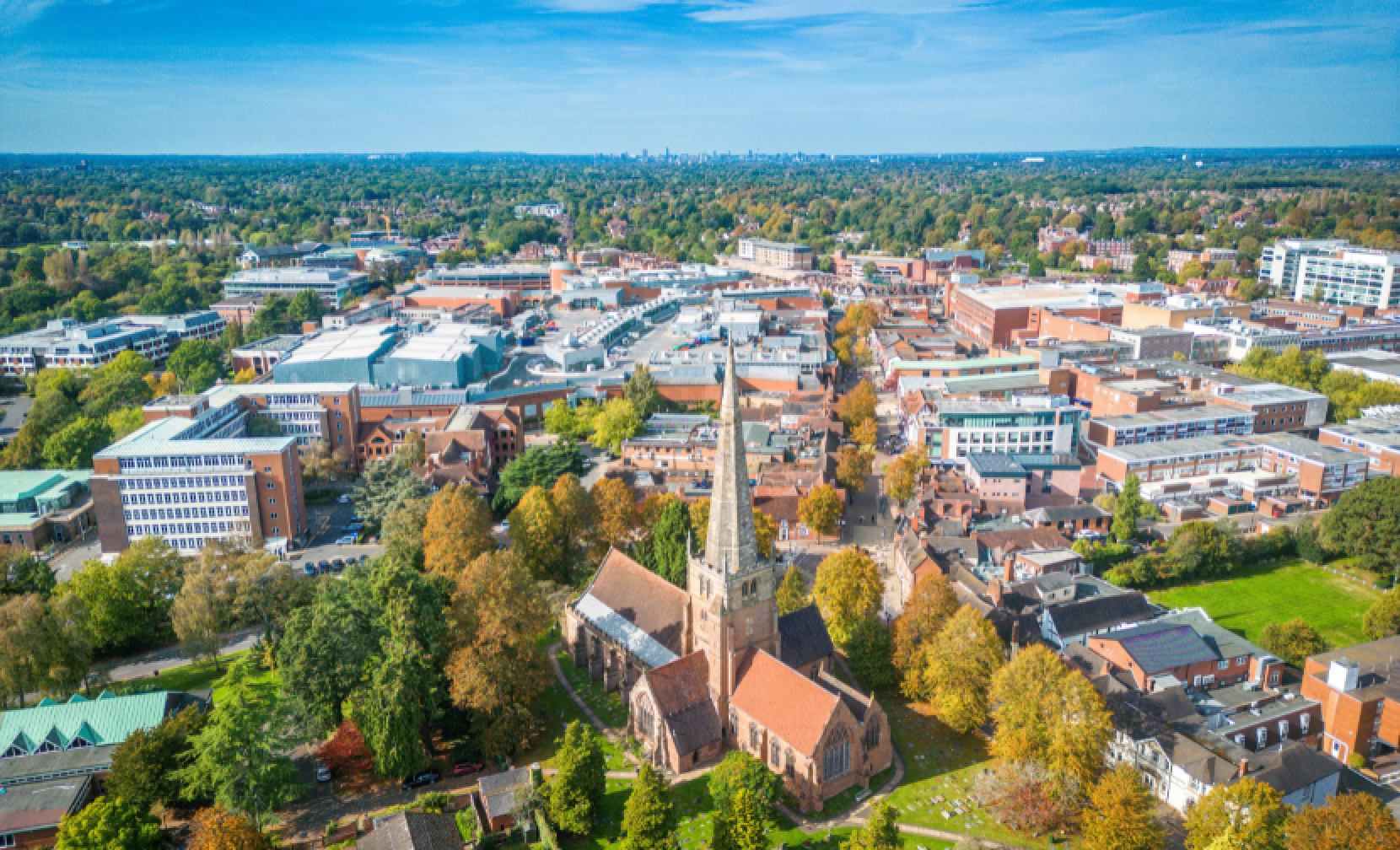 Aerial view of a suburban area featuring a church with a tall spire, surrounded by residential and commercial buildings, and lush green trees. The skyline of a city is visible in the distance under a clear blue sky.