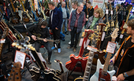 A crowded guitar show with various electric and acoustic guitars on display. A young man sits playing a guitar, while several attendees look around at the instruments on the stands. People are engaged in conversation, surrounded by an array of colorful guitars.