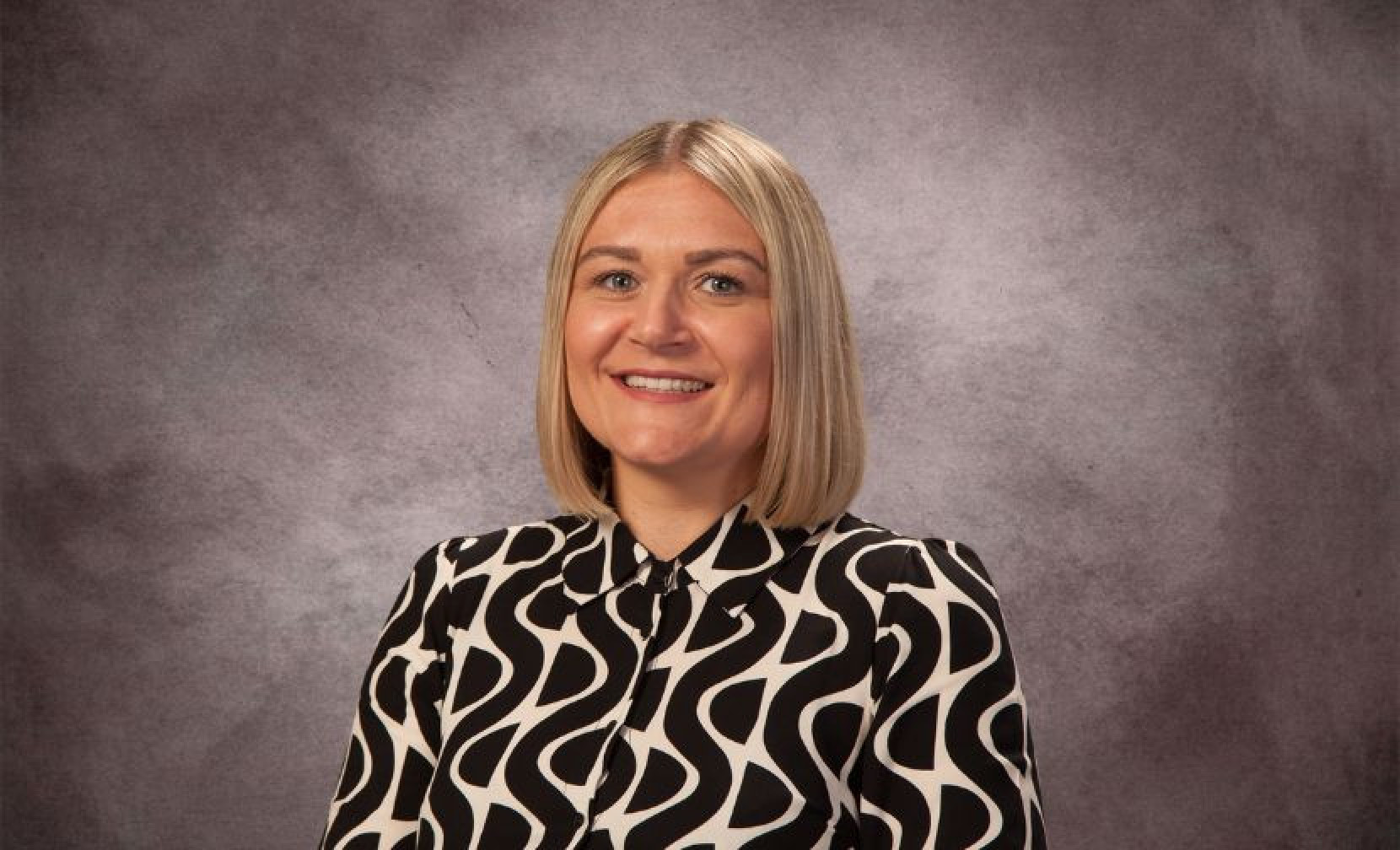 A woman with blonde hair styled in a bob wears a black and white patterned blouse. She smiles in front of a textured gray background.