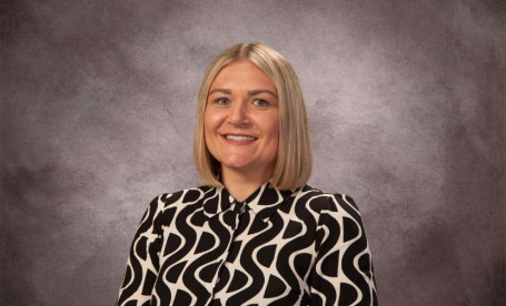 A woman with blonde hair styled in a bob wears a black and white patterned blouse. She smiles in front of a textured gray background.