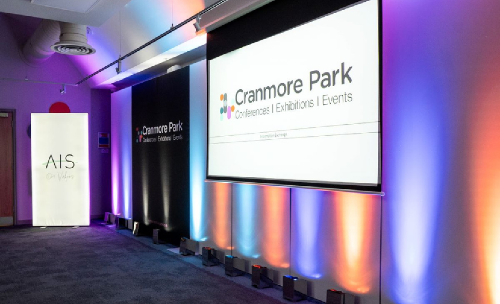 A well-lit conference room featuring colorful lighting and two displays. One screen shows the Cranmore Park logo, and a banner on the left displays the AIS logo.