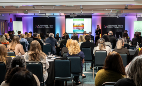 A conference room filled with attendees seated at tables, facing a speaker presenting on stage. There are colorful banners in the background, and a large screen displays presentation content. The audience appears engaged with the speaker.