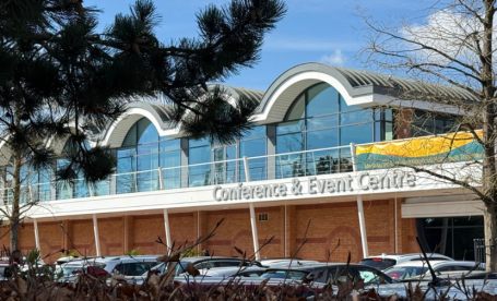 Modern conference and event center with large windows, a distinctive arched roof, and a banner across the front. Cars are parked in the vicinity under a clear blue sky.
