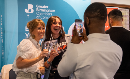 Two women smile while holding drinks, posing for a photo at a Greater Birmingham Chambers of Commerce event. One woman wears a 