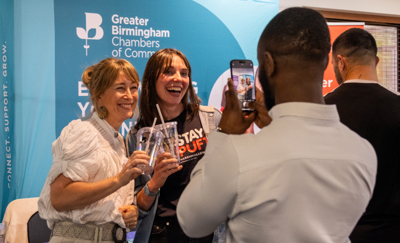 Two women smile while holding drinks, posing for a photo at a Greater Birmingham Chambers of Commerce event. One woman wears a 