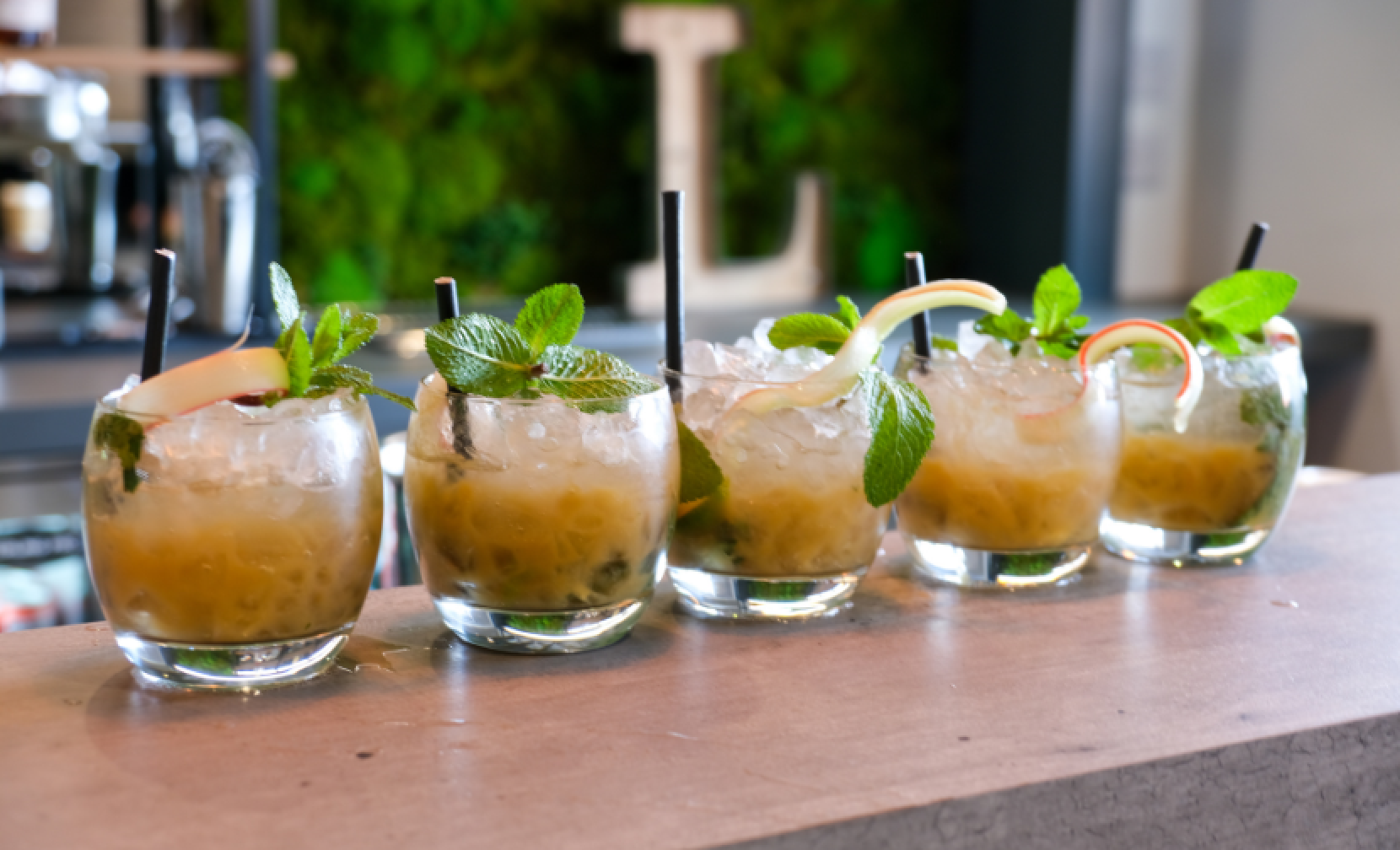 Five cocktail glasses featuring mint springs, black straws, crushed ice, and apple curls, are lined up in a row on top of a bar counter. A wooden letter L for 'The Loft' is just visible in the background against a green living wall.
