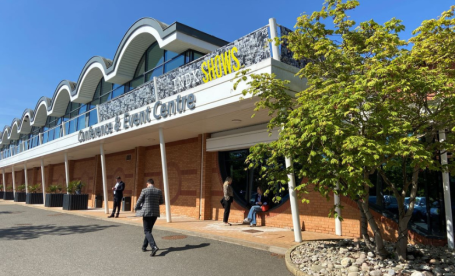A modern event center with a wavy roof design and large windows. People walk in front of the building, which features a sign that reads 