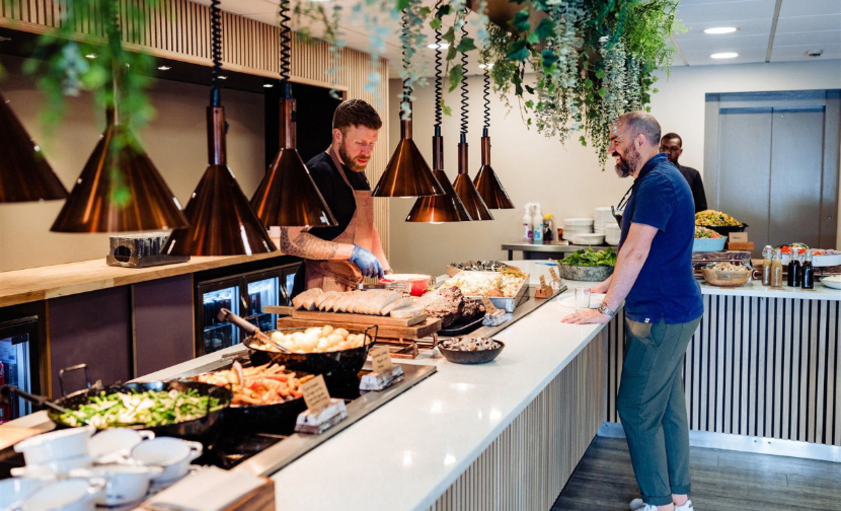 A modern food station featuring a chef preparing dishes while a customer observes. The counter is filled with various appetizers and salads, surrounded by stylish pendant lights and greenery.