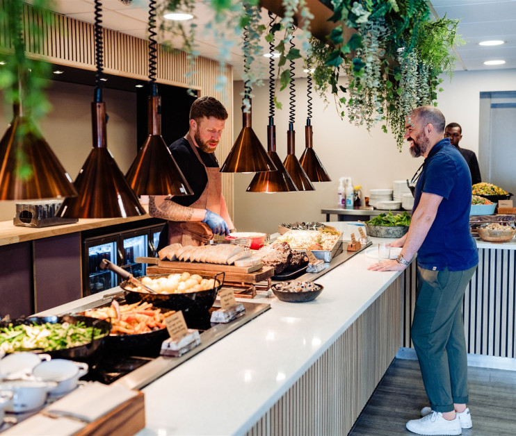 A buffet-style food counter with a chef preparing dishes while a customer watches. The counter features various platters of food, including salads and roasted items, under pendant lighting and surrounded by greenery.