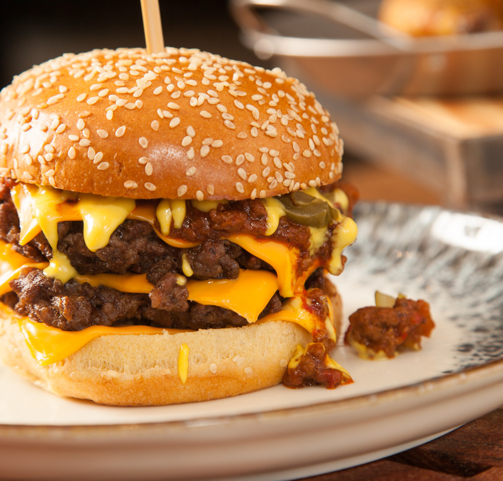 A towering cheeseburger stacked with two juicy patties, melted cheddar cheese, and topped with spicy jalapeños and a rich sauce, served on a sesame seed bun. A side of rustic fries is blurred in the background.