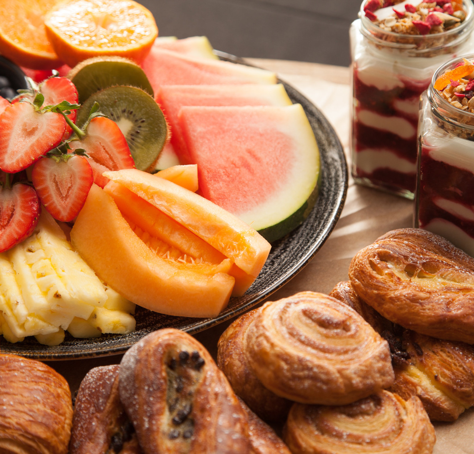 A colorful fruit platter featuring slices of watermelon, cantaloupe, honeydew, strawberries, kiwi, and oranges, alongside two jars of yogurt parfaits topped with granola and dried fruits. Fresh pastries, including croissants and cinnamon rolls, are also arranged nearby.