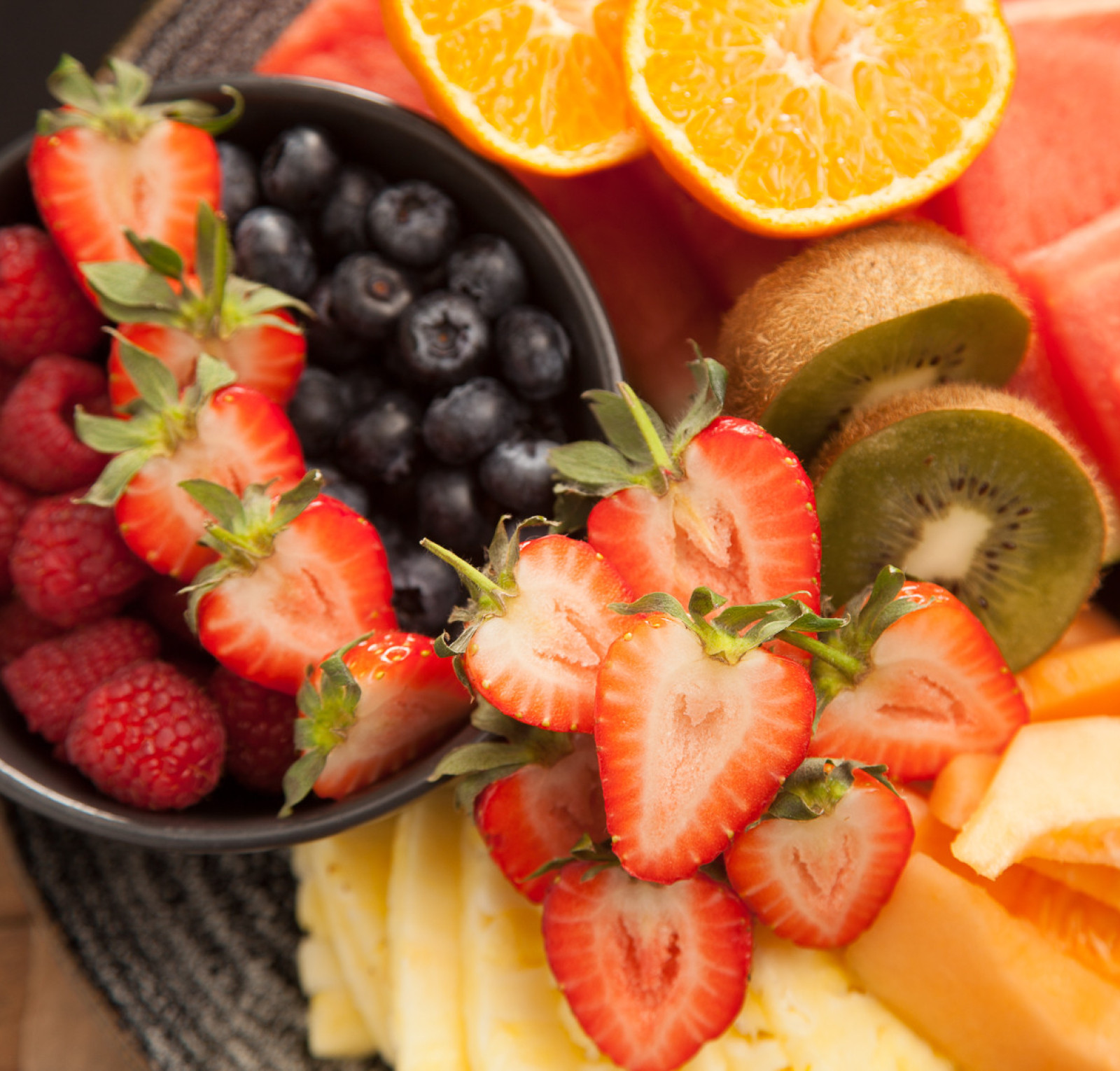 A vibrant fruit platter featuring a variety of fresh fruits. There's a bowl filled with blueberries and raspberries, surrounded by sliced strawberries, kiwis, watermelon, cantaloupe, pineapple, and orange segments.
