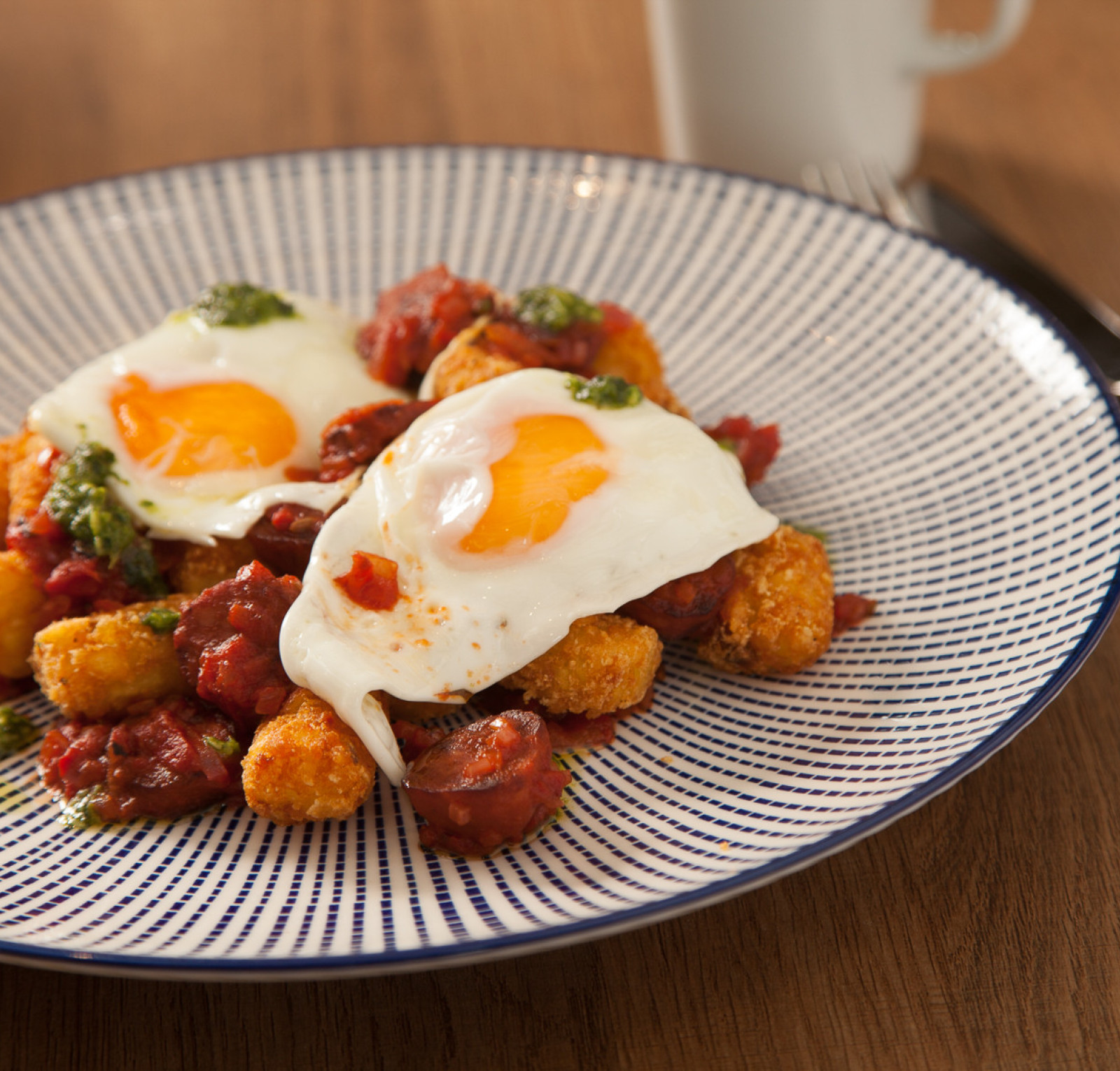 A plate of crispy tater tots topped with chorizo and two sunny-side-up eggs, garnished with a drizzle of green sauce and served on a decorative blue and white patterned plate.