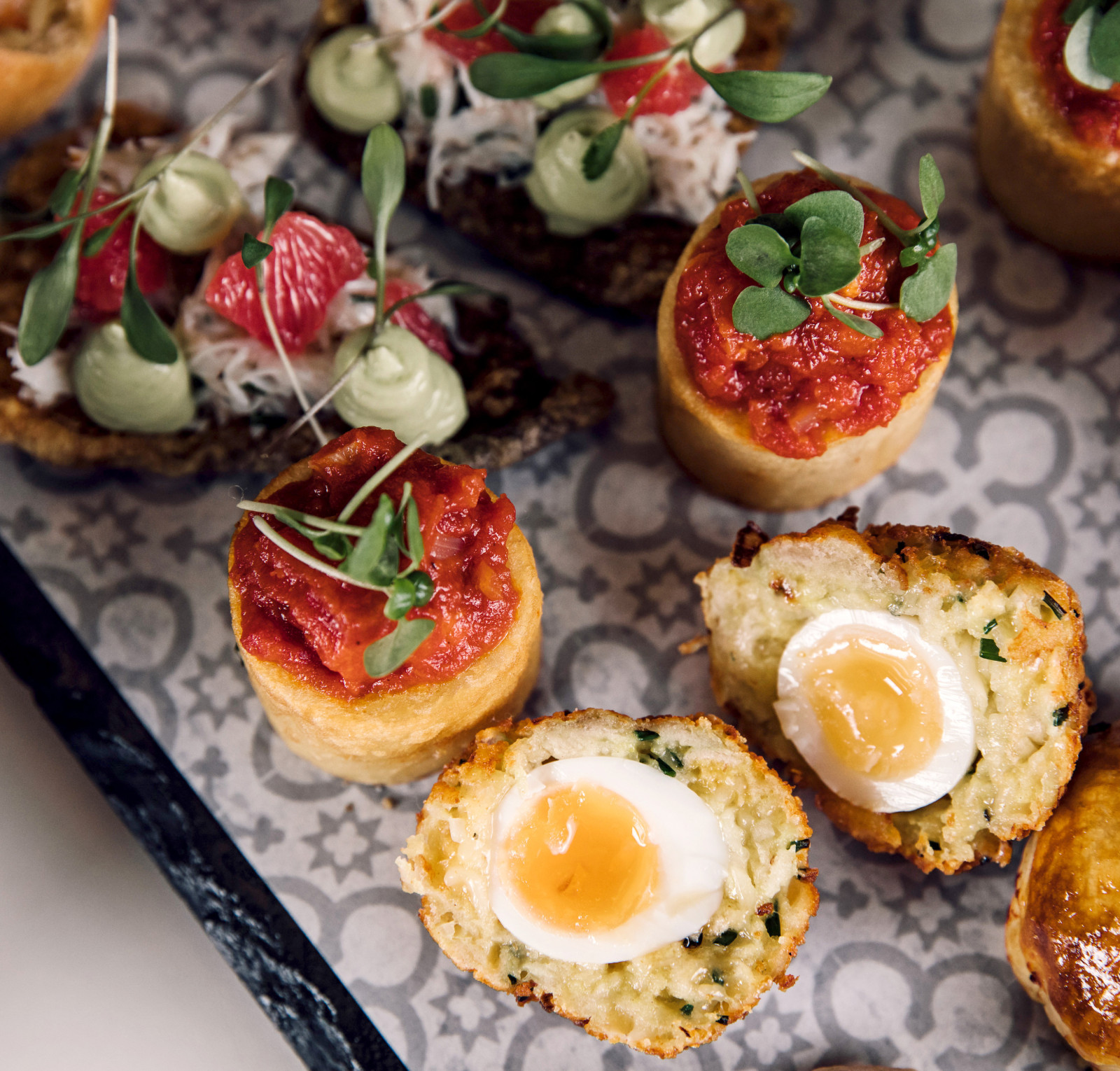 A platter of gourmet appetizers featuring various bite-sized treats. Some are topped with vibrant sauces and garnishes, while others have soft-boiled eggs nestled within crumbly bases. The arrangement is visually appealing with fresh herbs adding a pop of color.