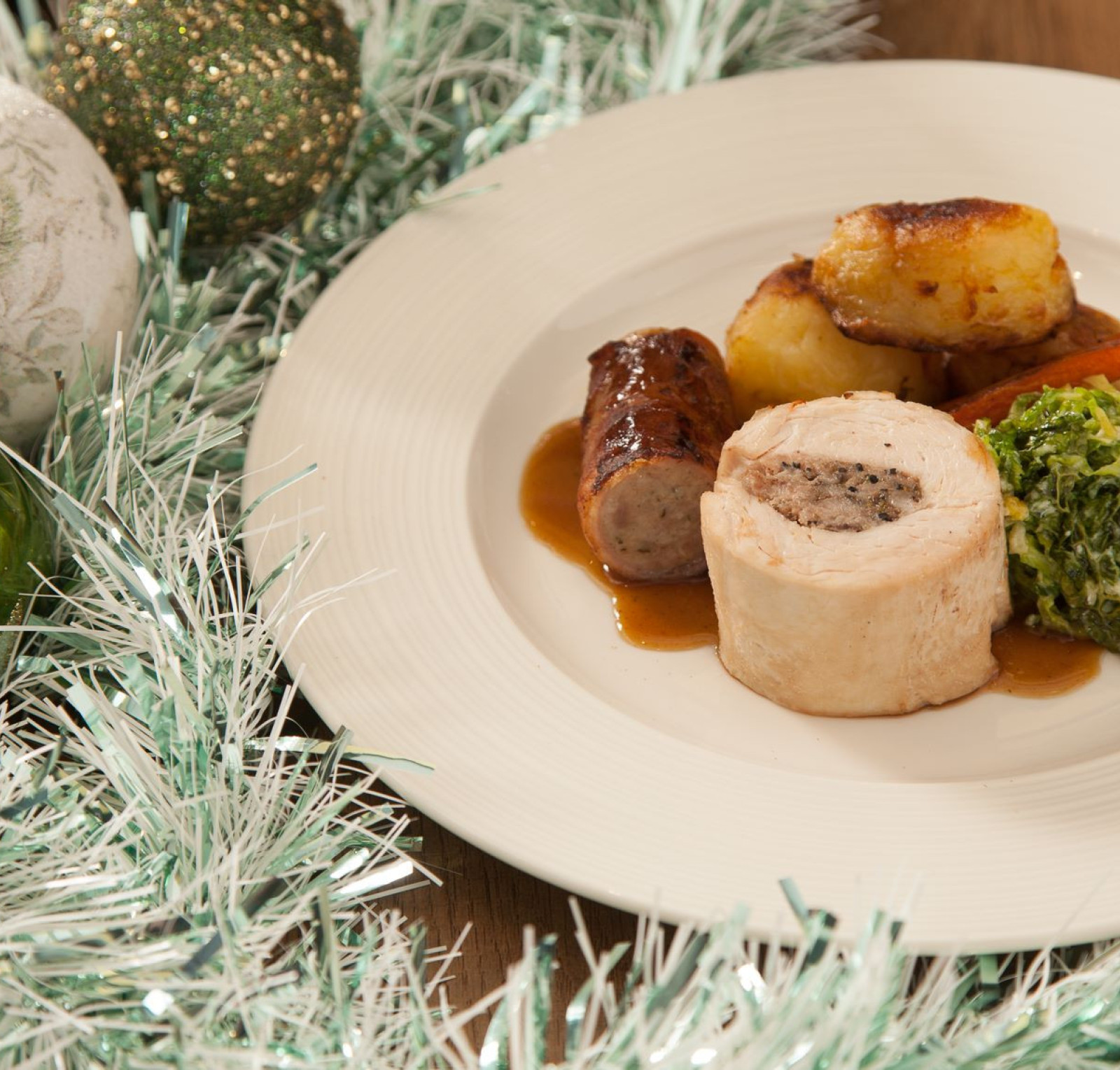 A festive plate featuring a slice of stuffed poultry, accompanying sausages, roasted potatoes, sautéed greens, and a drizzle of sauce. Decorated with sparkling green and silver ornaments and festive tinsel.