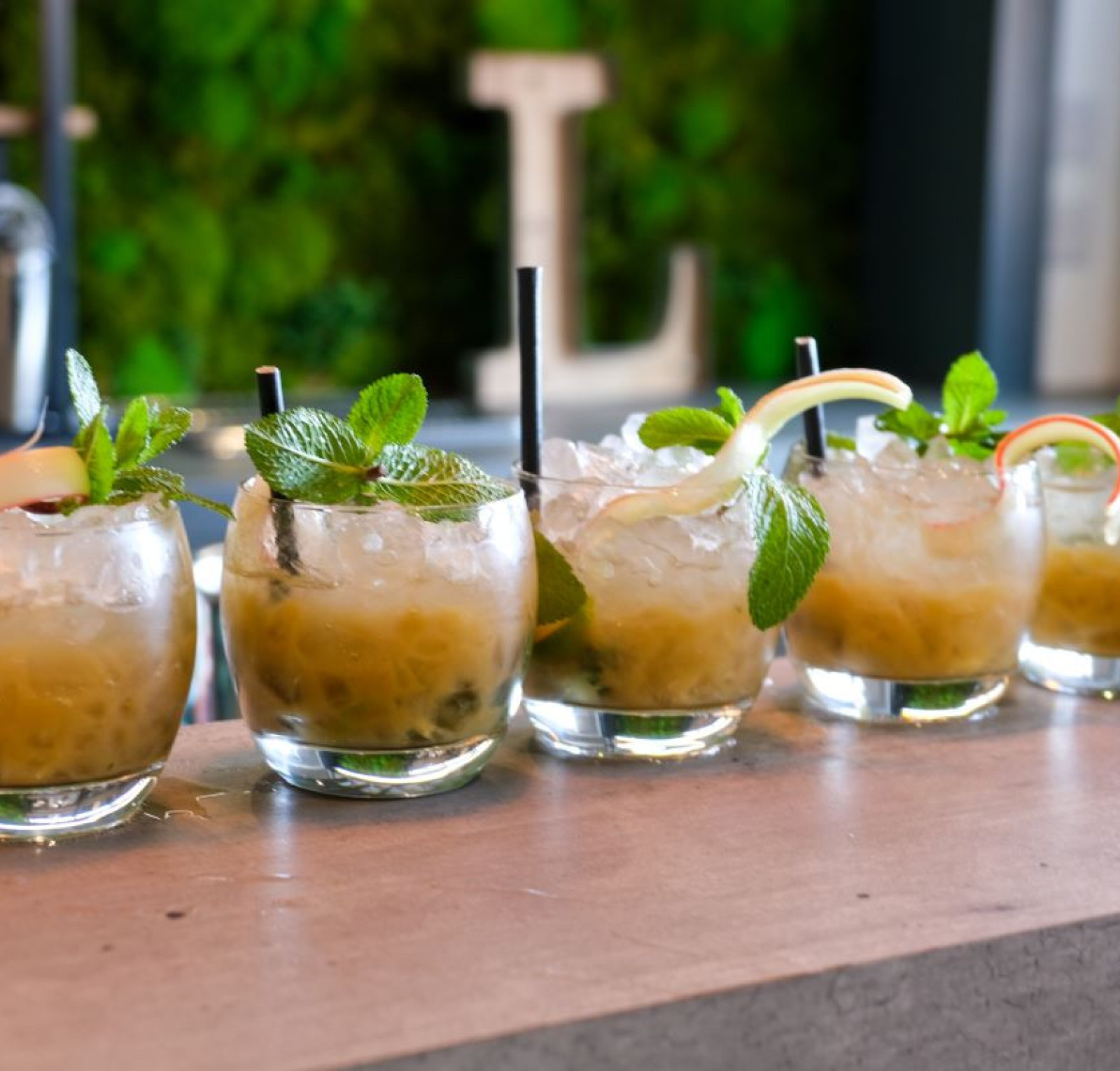 A row of six cocktails garnished with mint leaves and sliced fruit. Each drink is served in a clear glass filled with ice and a straw, set against a green moss backdrop.