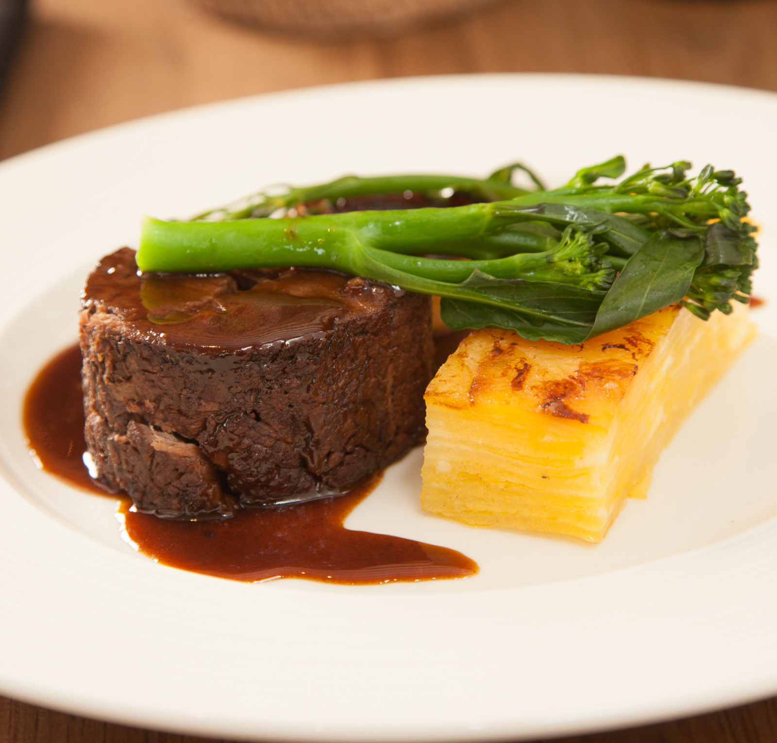 A gourmet dish featuring a round slice of tender beef, topped with rich brown sauce, accompanied by a portion of golden, layered potato and vibrant green broccoli stems.