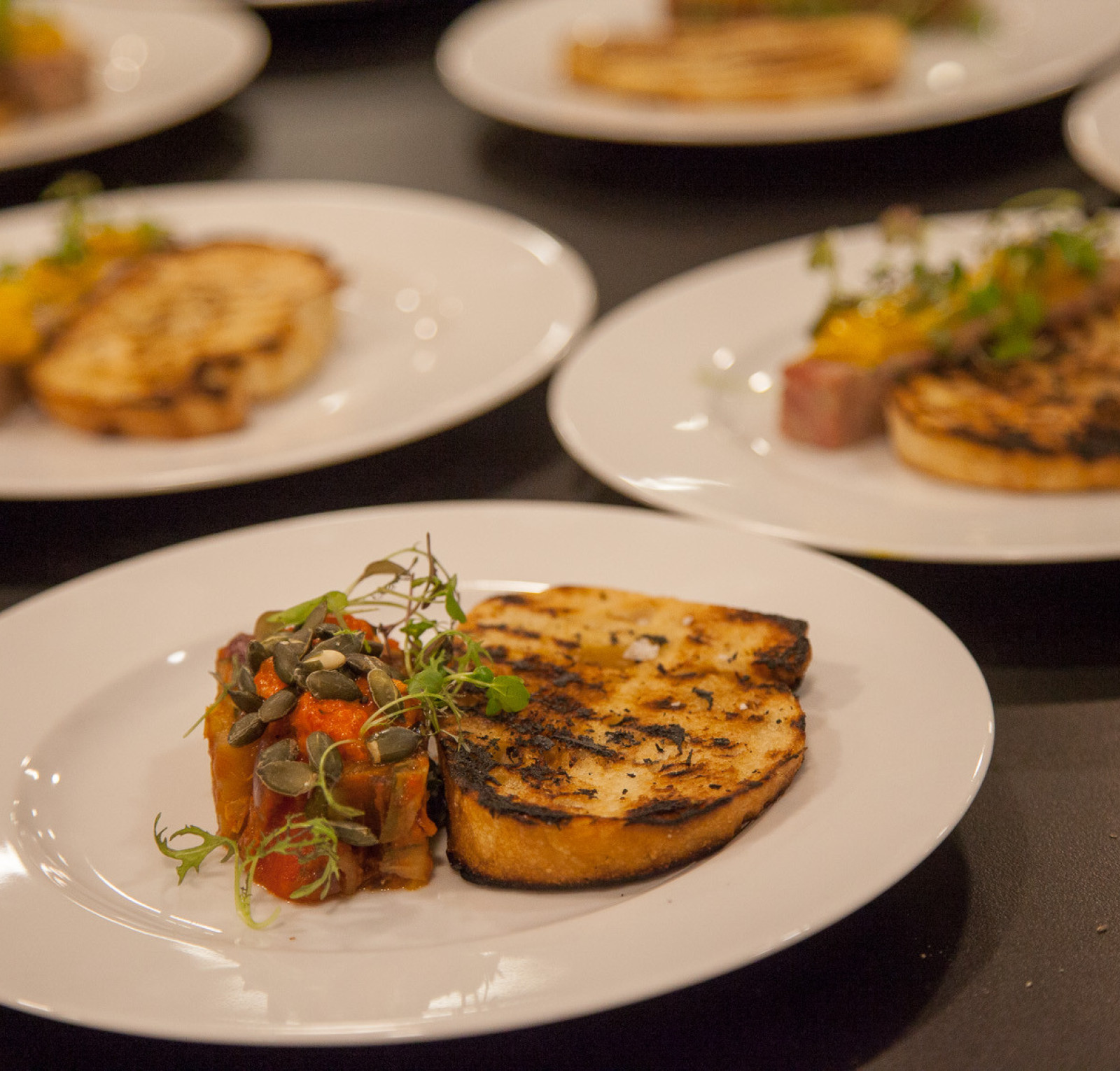 A white plate features a grilled slice of bread alongside a colorful vegetable medley topped with microgreens. Additional plates in the background display different grilled dishes.