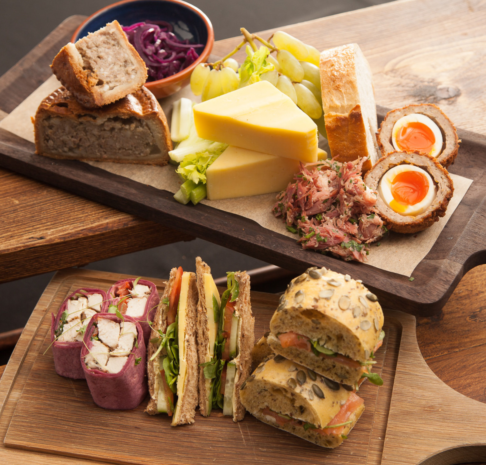 A wooden serving board displays a variety of foods including slices of cheese, pickled red cabbage, green grapes, a meat pie, scotch eggs, and a mixture of sandwiches with colorful fillings.