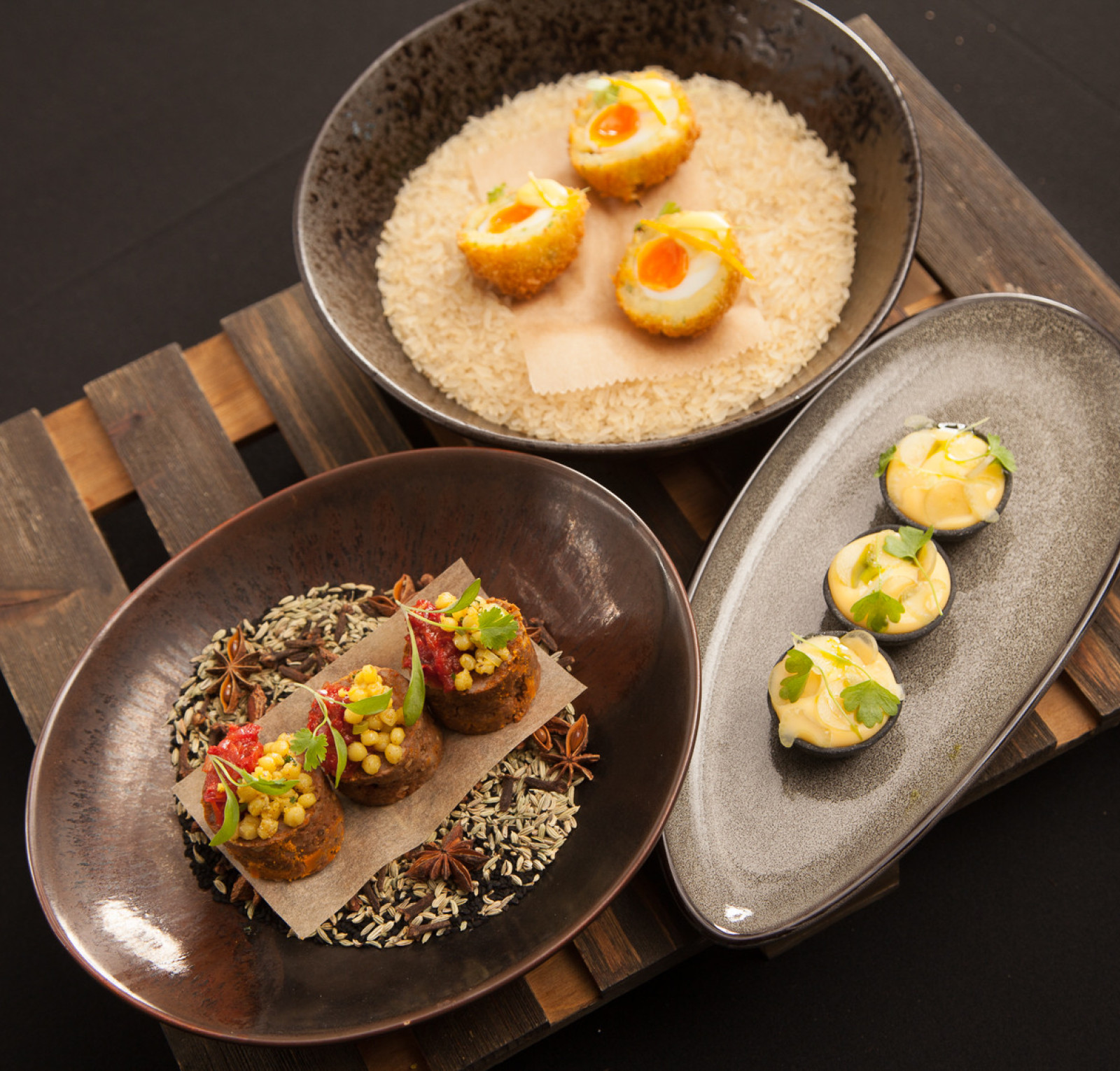 A beautifully arranged dish featuring three different types of appetizers. The first plate contains golden fried balls garnished with a vibrant sauce, served on a bed of rice. The second plate has colorful bite-sized rolls topped with vegetables and sauces, arranged neatly. The third plate includes small, round desserts adorned with fresh herbs, all presented on rustic wooden surfaces.
