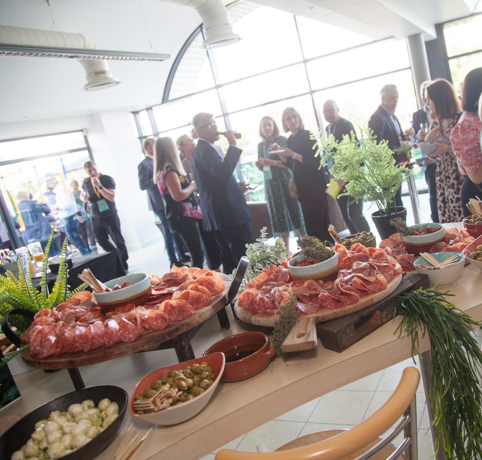 A well-decorated event space featuring guests mingling and enjoying refreshments. A display of charcuterie, including meats and olives, is set on a table in the foreground, complemented by greenery.