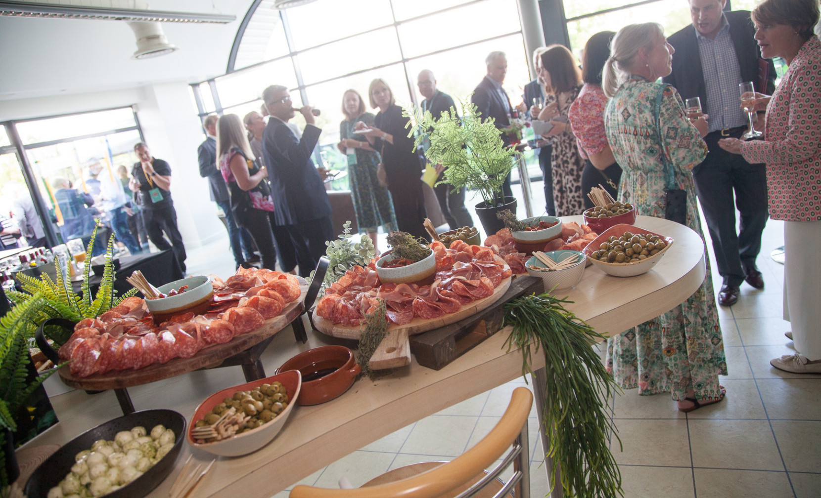 A well-decorated event space featuring guests mingling and enjoying refreshments. A display of charcuterie, including meats and olives, is set on a table in the foreground, complemented by greenery.