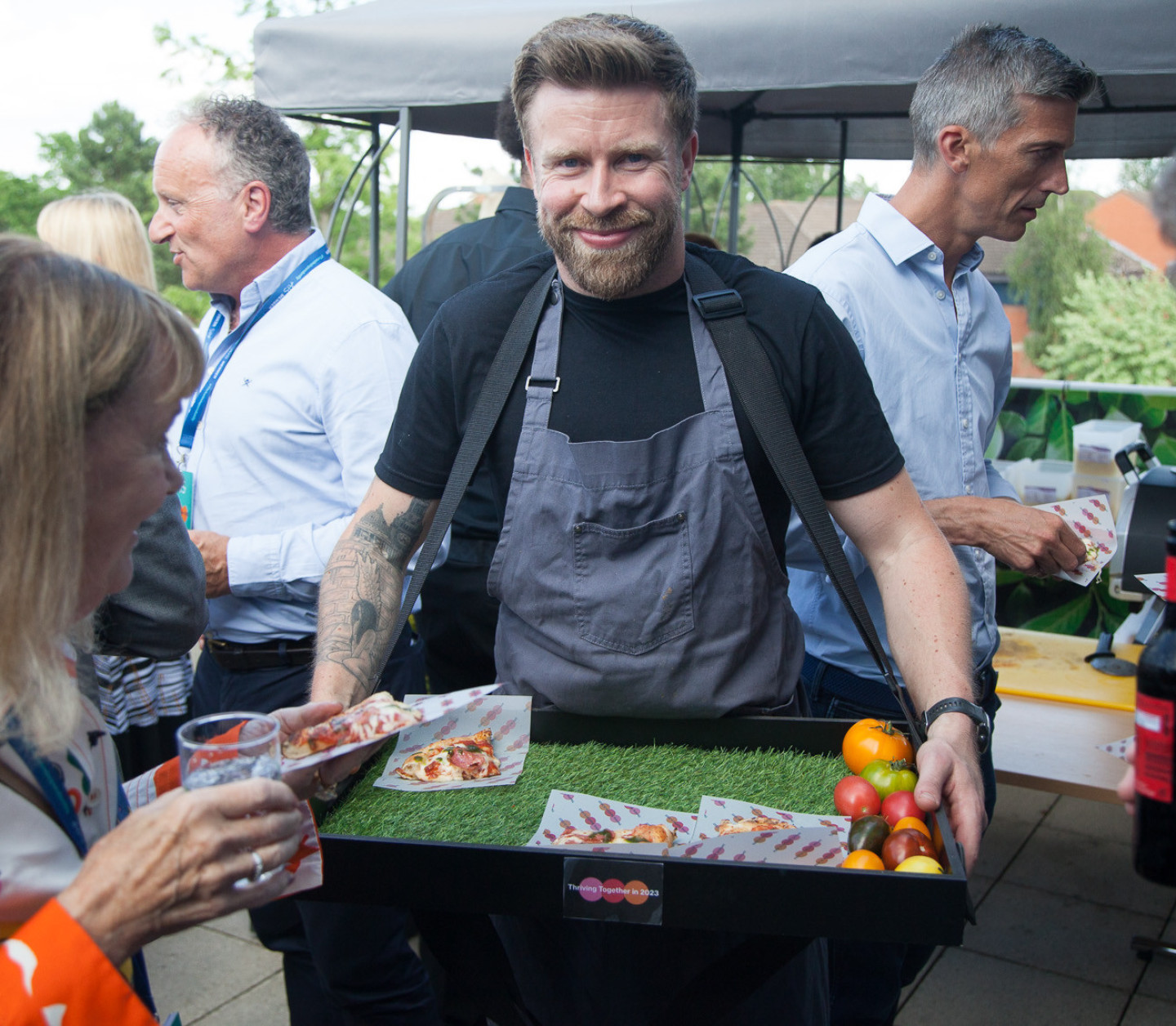 A spacious dining area featuring multiple wooden tables and chairs in various styles. The room is well-lit, with large windows and plants adding greenery. A buffet area is visible in the background, and modern decor enhances the ambiance.