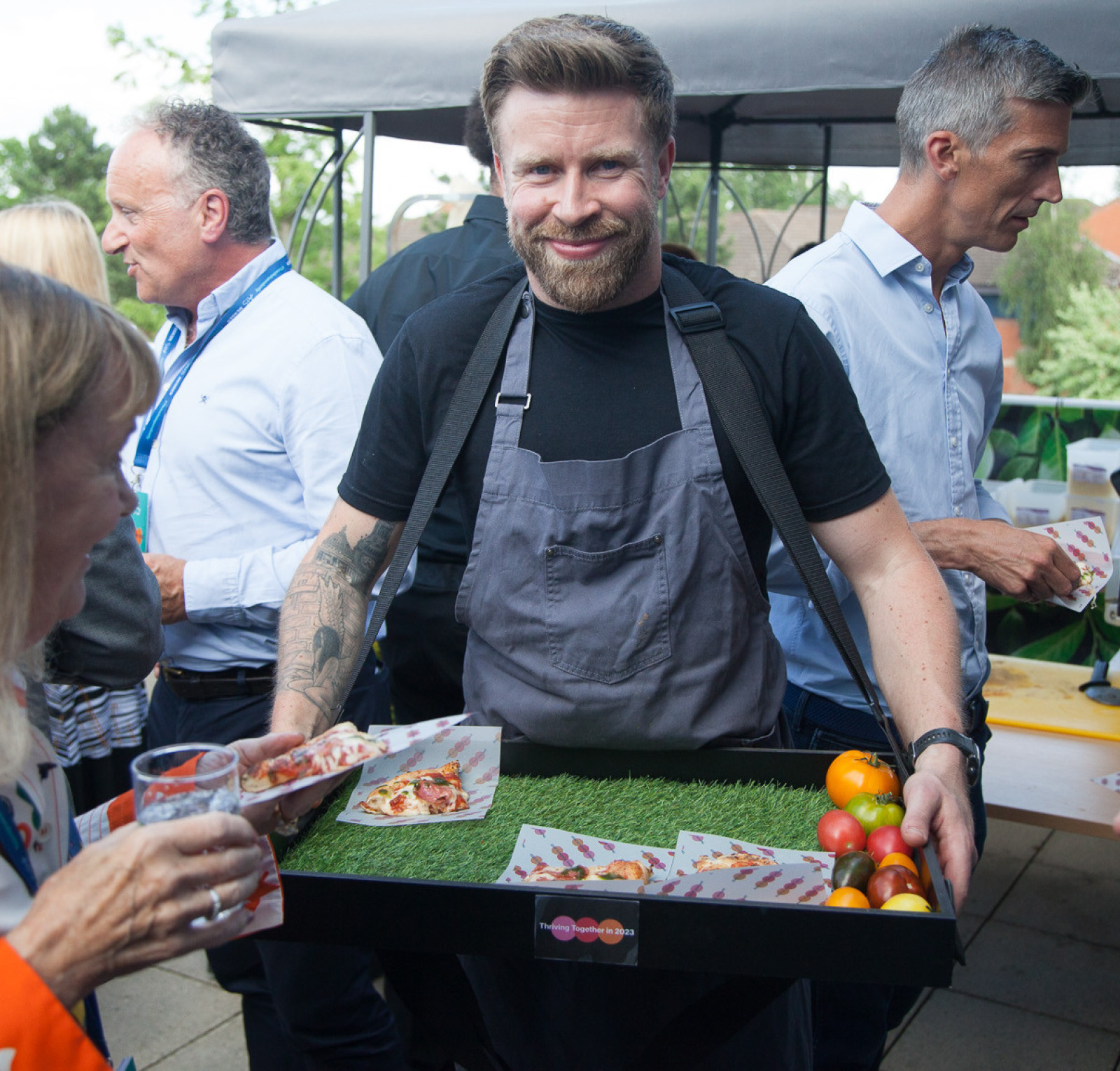 A man with a beard and tattoos wears an apron and smiles while holding a tray of food, including pizza slices and fruits, at an outdoor event with attendees mingling around him.