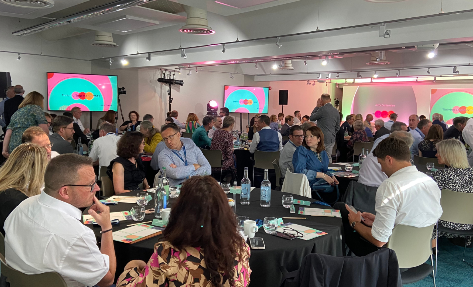 A spacious room filled with attendees seated at tables, engaged in discussions. Several screens display colorful slides, and there are various setups for presentations and lighting. The atmosphere appears lively and collaborative.