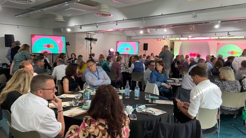 A spacious room filled with attendees seated at tables, engaged in discussions. Several screens display colorful slides, and there are various setups for presentations and lighting. The atmosphere appears lively and collaborative.