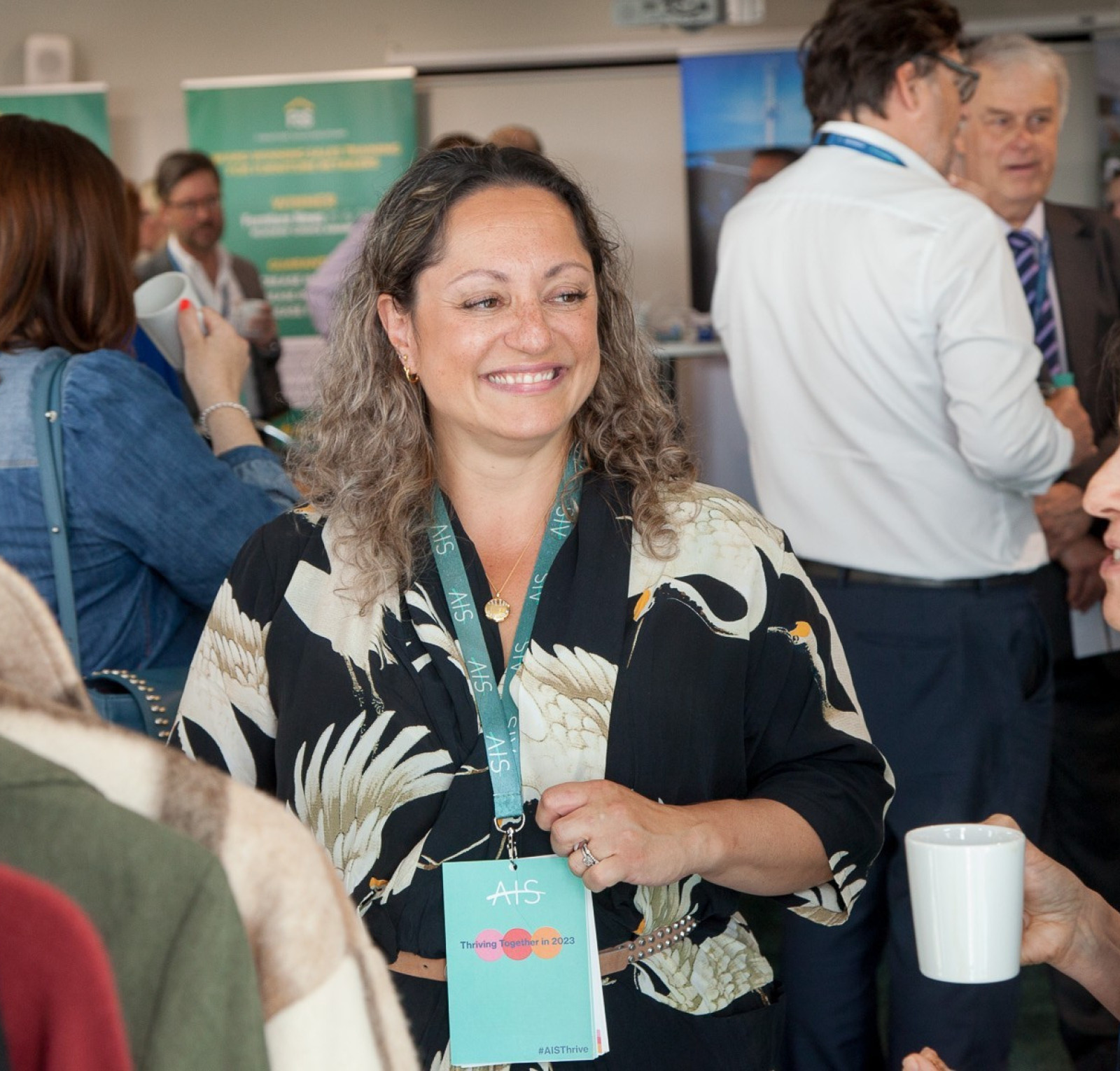 A woman with curly hair wearing a floral shirt smiles while holding a cup, engaging in conversation at a networking event. People are mingling in the background, with various banners and displays visible.