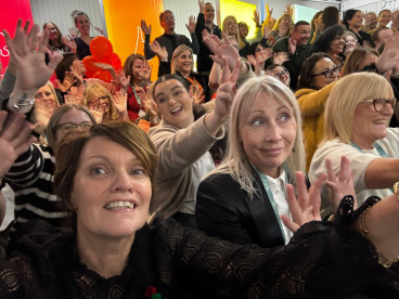 A large group of people are enthusiastically waving their hands and smiling while gathered together in a lively setting, with colorful background displays.