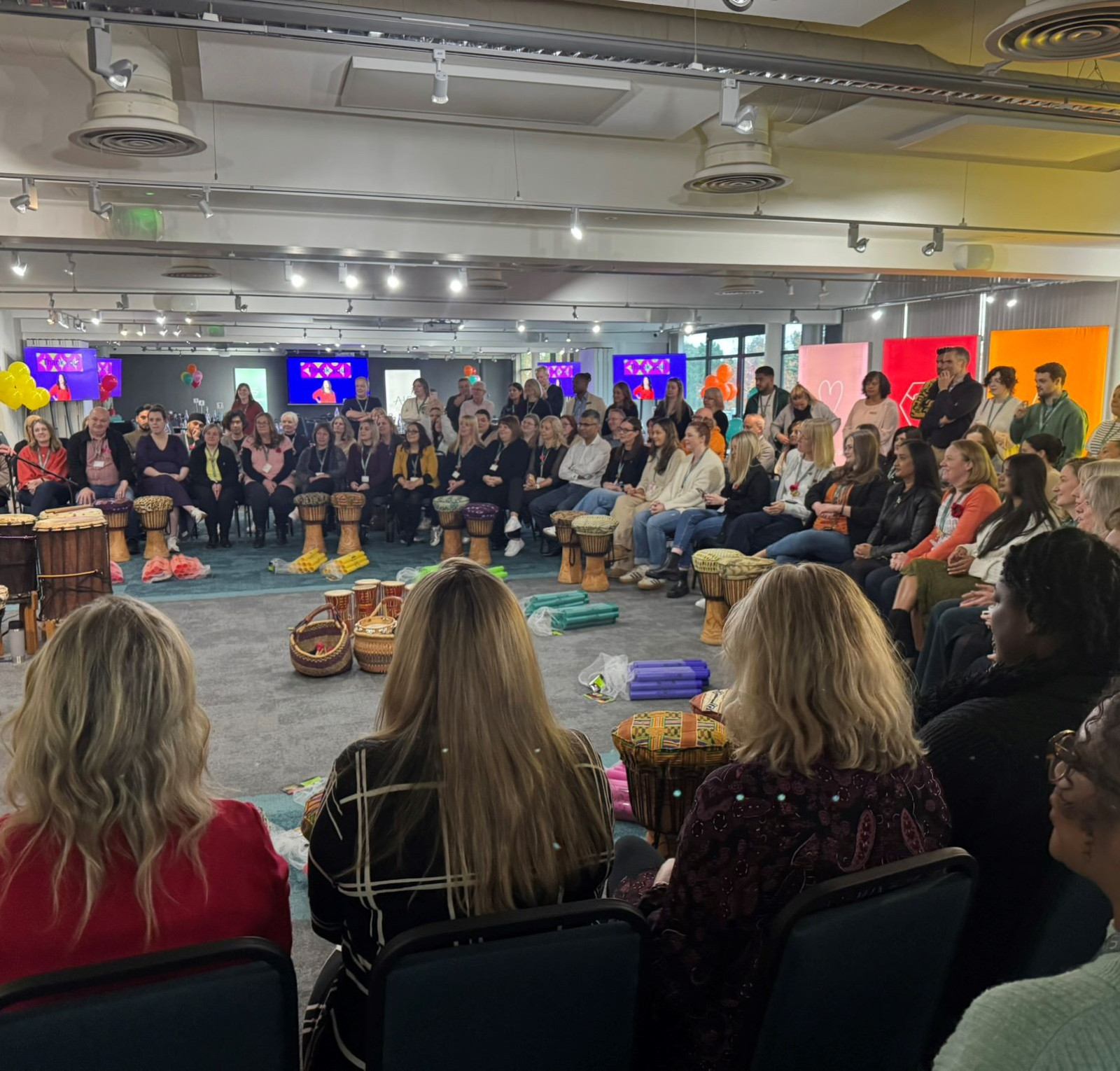 A large group of people seated in a circle, participating in a drumming session in a bright and modern room. Colorful decorations and screens are visible in the background.