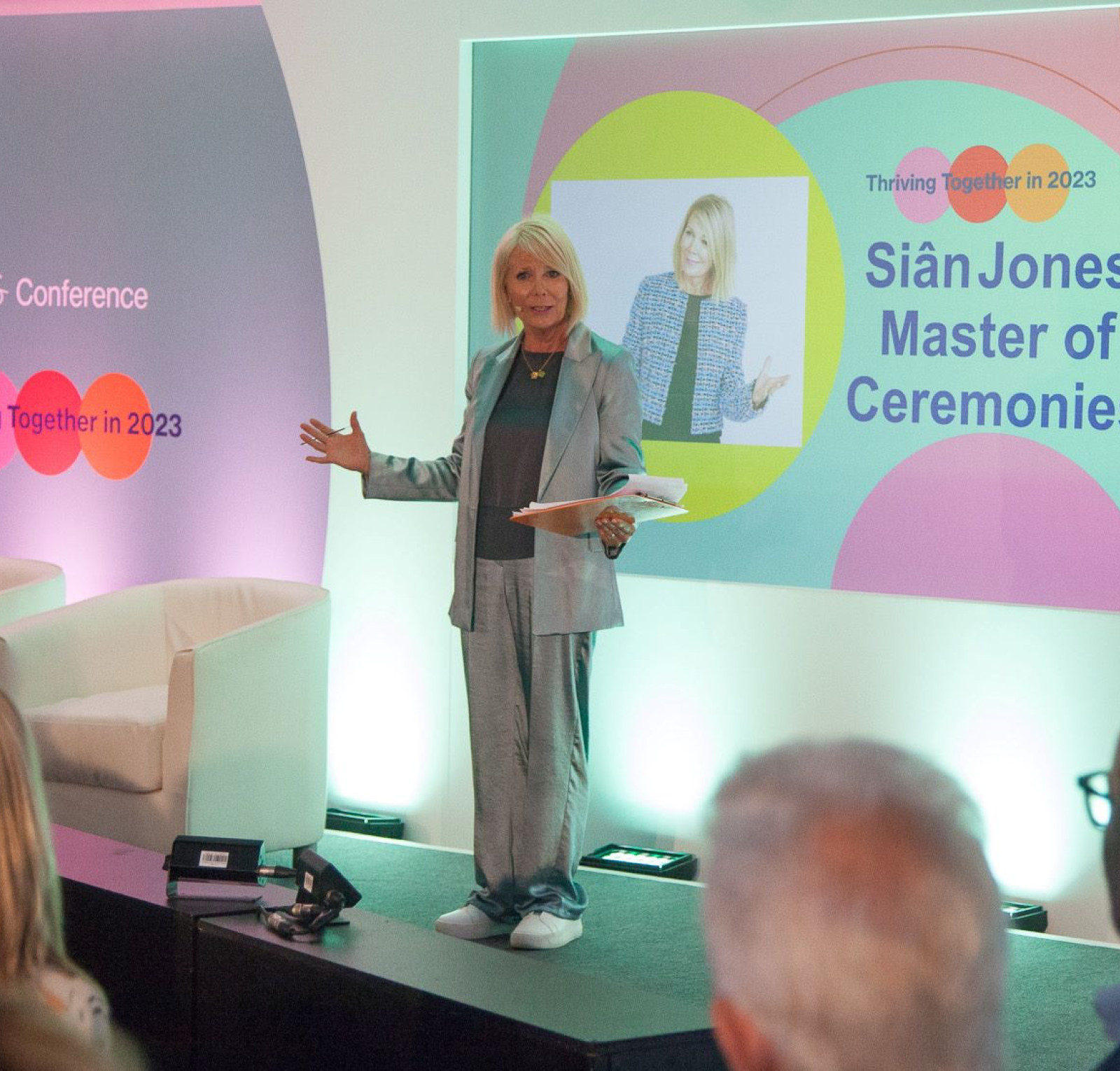 Sian Jones stands on stage as the Master of Ceremonies at the ATS Conference, engaging the audience with a smile. The backdrop features colorful graphics with the text 