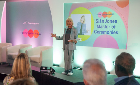 Sian Jones stands on stage as the Master of Ceremonies at the ATS Conference, engaging the audience with a smile. The backdrop features colorful graphics with the text 