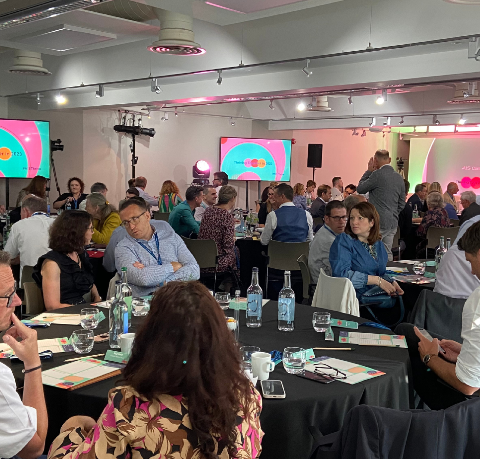 A large group of people seated and engaged in discussions at a conference or event. Colorful presentation slides are visible on screens in the background. Tables are set with notebooks, drinks, and materials for attendees.