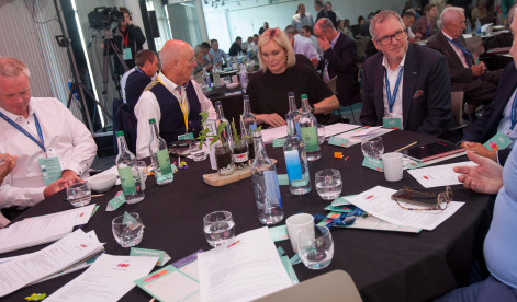 A group of professionals engaged in discussion around a conference table, with glasses of water and documentation scattered across the table. Some participants are smiling and interacting, while others are focused on the conversation. The setting appears to be a modern conference room with bright lighting.