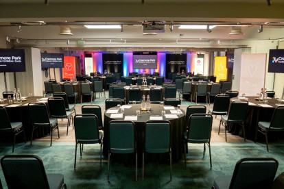 A conference room set up with multiple rows of tables and chairs, featuring branding banners along the walls. Two screens are displayed at the front, and the lighting creates a vibrant atmosphere.