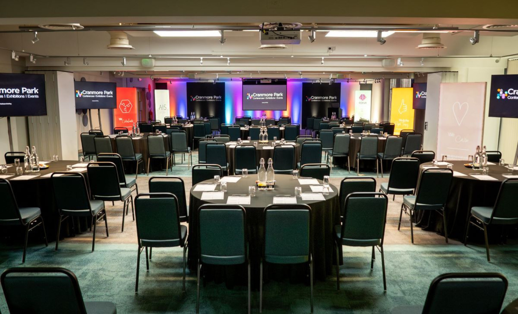 A conference room set up with multiple rows of tables and chairs, featuring branding banners along the walls. Two screens are displayed at the front, and the lighting creates a vibrant atmosphere.