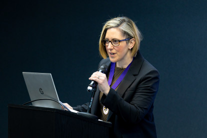 A woman with blonde hair, wearing glasses and a black suit, speaks into a microphone while standing at a podium with a laptop in front of her. She appears to be giving a presentation.