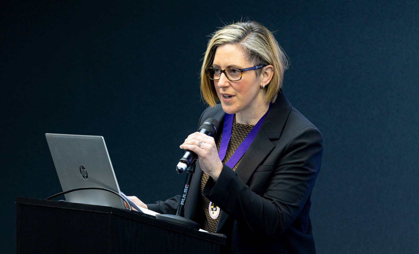 A woman with blonde hair, wearing glasses and a black suit, speaks into a microphone while standing at a podium with a laptop in front of her. She appears to be giving a presentation.