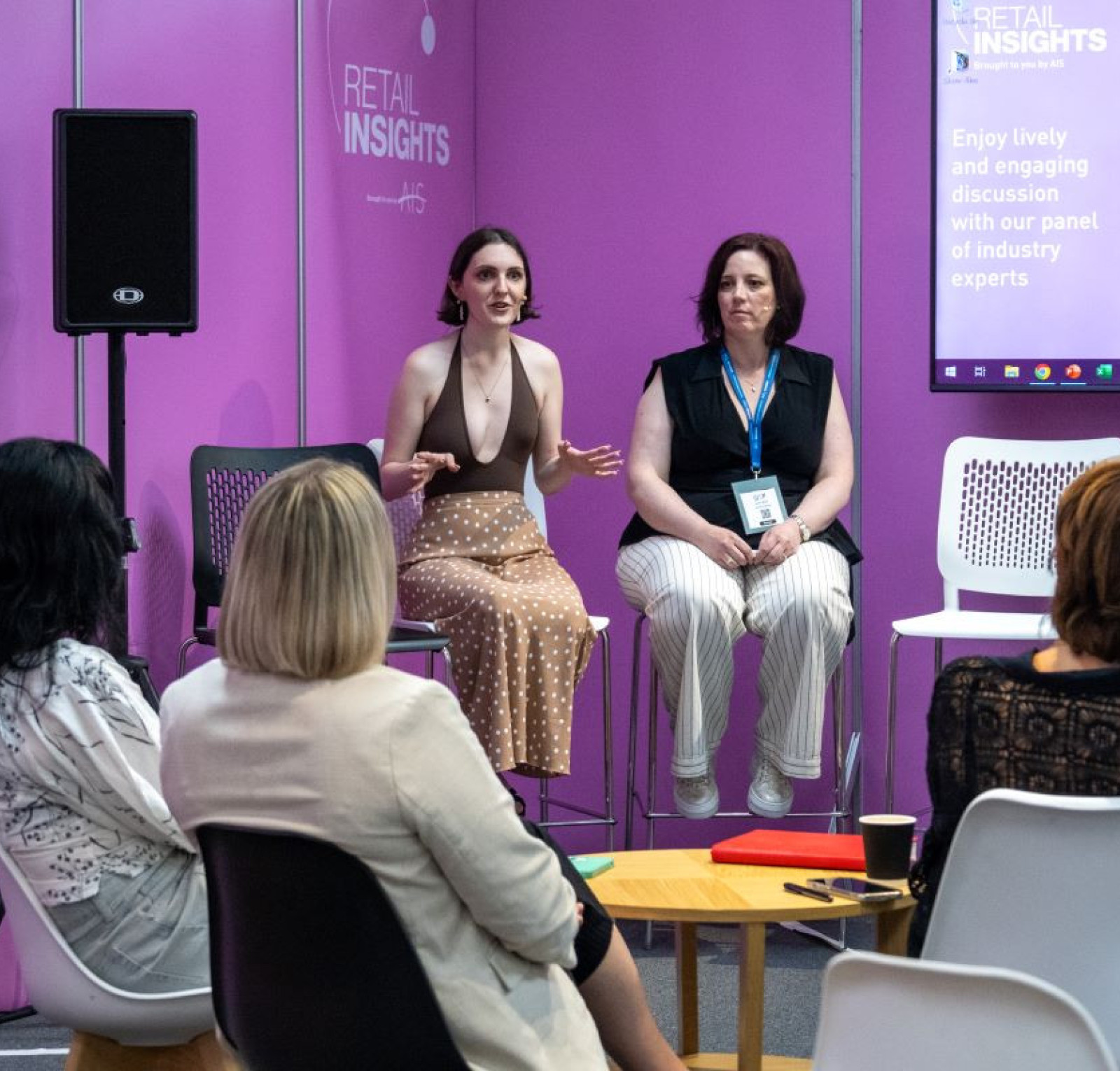 Two women are sitting on a stage, engaging in a discussion at a Retail Insights event. One woman is speaking animatedly while the other listens attentively. In the foreground, a small audience is seated, focused on the speakers. The backdrop features a vibrant pink wall with text related to the event.